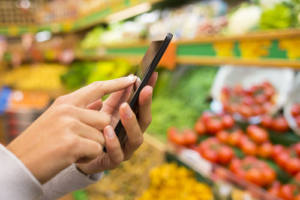 Woman,Using,Mobile,Phone,While,Shopping,In,Supermarket,,Vegetable,Department